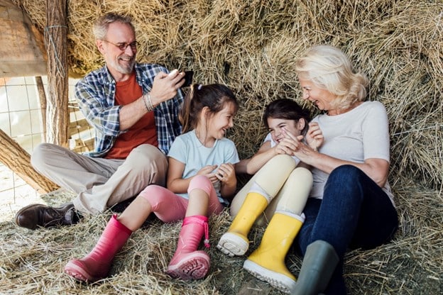 Grandfather taking image of wife and grandchildren with cellphone while sitting on hay.