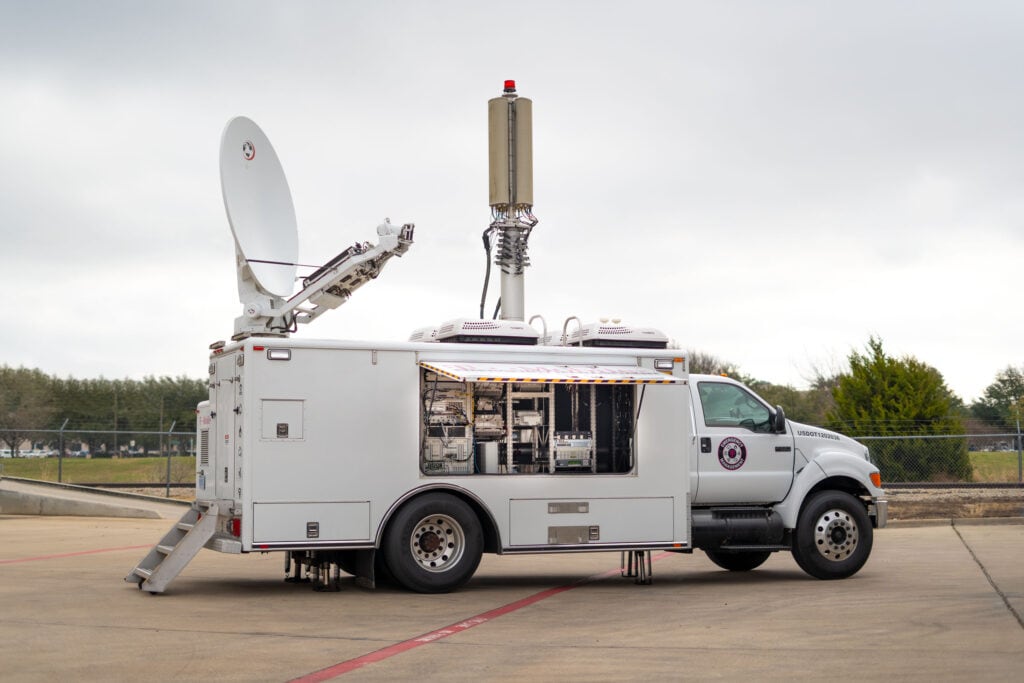 Large truck with mounted satellite dish and antenna tower. Vehicle is branded with the mobile365 Emergency Management logo and has an open window to display the interior equipment.